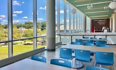 Rows of tables with blue chairs are next to large windows with a sign in the back reading “Welcome to the Daniel and Grace Tully Dining Commons, The Tully”