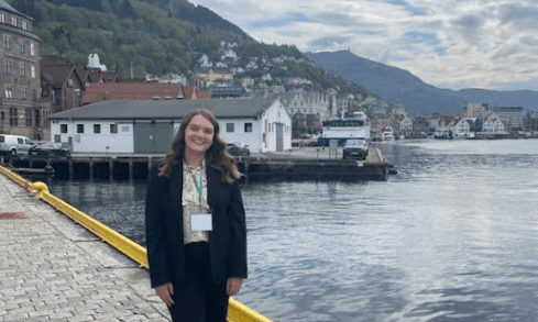 A student wearing a lanyard poses in front of a Norwegian harbor.