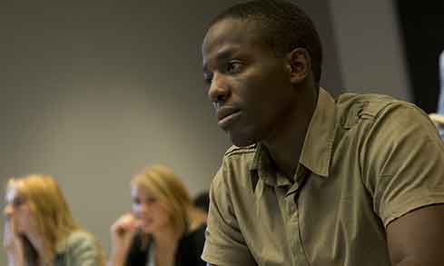 A person in a khaki shirt attentively listens in a classroom setting, with others sitting in the background.