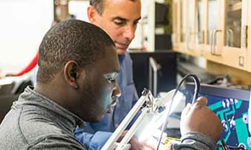 Two men collaborating on a computer in a laboratory setting, focused on their work and surrounded by scientific equipment.