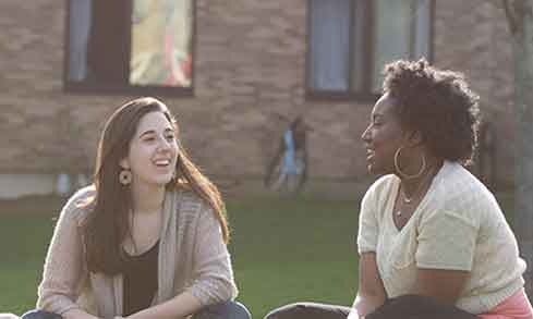 Two women seated on the grass engaged in a conversation, enjoying a sunny day outdoors.