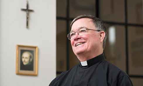 A priest stands in front of a cross and a painting. 