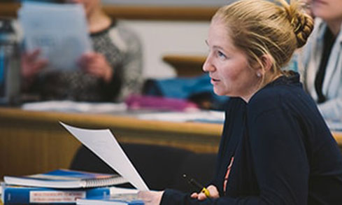 A woman sits at a desk in a classroom, surrounded by papers and books.