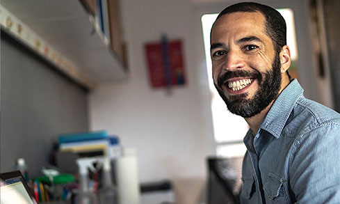 A smiling man with a beard sits at a desk.