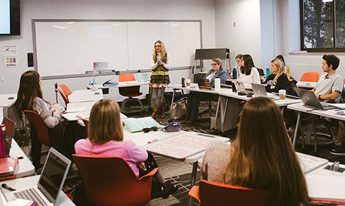 Students sit at desks in a classroom, listening attentively to their teacher at the front of the room.