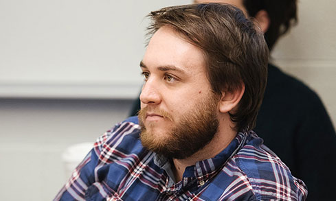 A bearded man in a plaid shirt sits in a classroom, focused on the lesson.