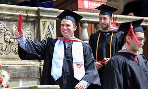 Student in cap and gown walking down steps holding out diploma baton after graduating at a commencement ceremony.