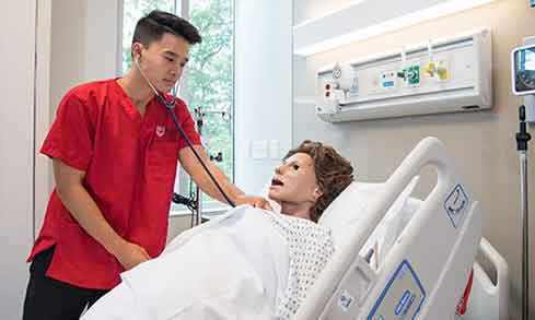 Student wearing red Fairfield scrub using a stethoscope on a dummy. 