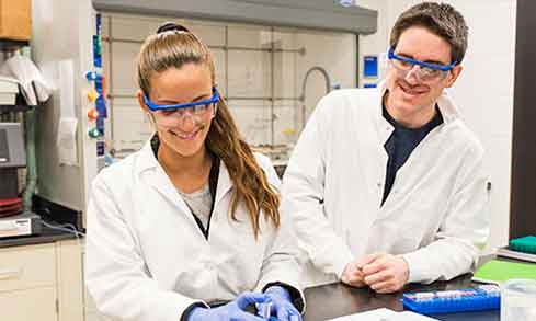 Two students wearing lab coats and eye protection in the school lab. 