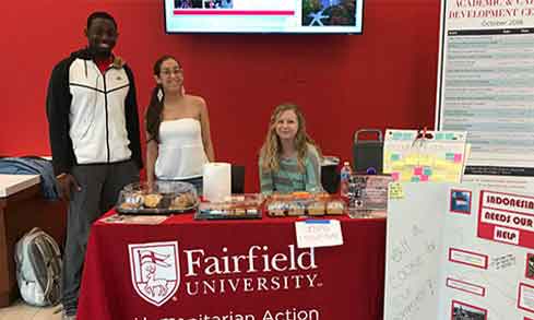 three students sit behing a table with a red tablecloth that says humanitarian action.