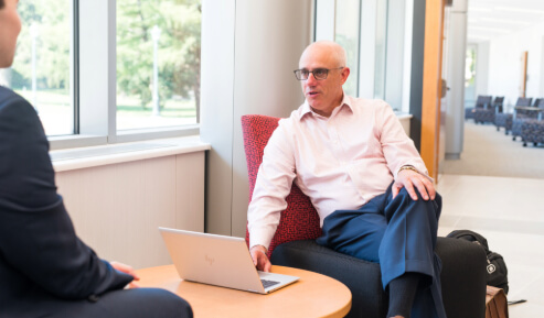 A professor sits in a chair while speaking to a student.