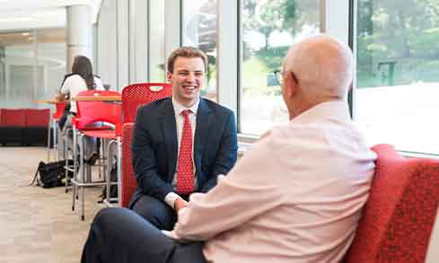 Two men in business suits engaged in conversation while seated in a modern lobby setting.