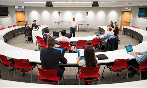 Students wearing formal attire listening to a professor give a lecture in a circular classroom.