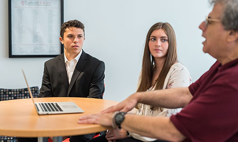 Students wearing formal attire listen to a professor.