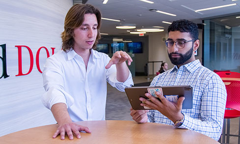 Two male students stand by a table, looking at a tablet together, engaged in conversation.