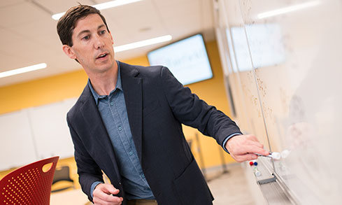 A professor in a suit stands confidently in front of a whiteboard, ready to present his ideas.