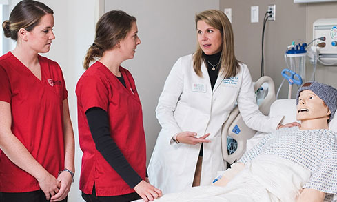 2 nursing students listen to professor who is working with manikin.