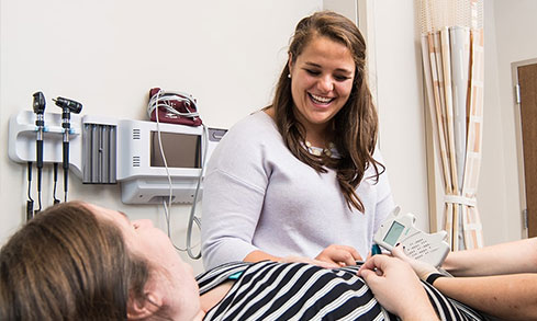 2 smiling nursing students stand over mock patient in doctor's office.