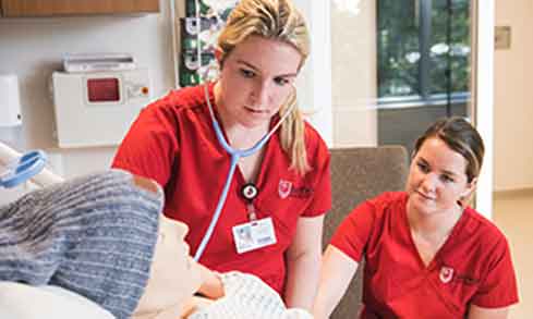 Two students in red scrubs use a stethoscope on a manikin.