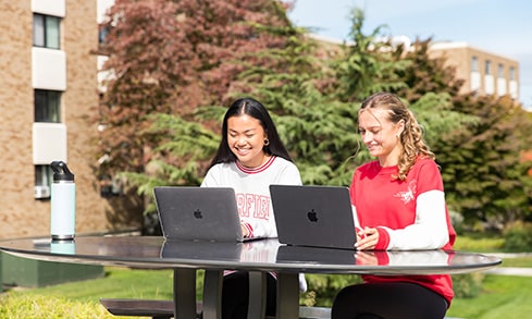 Two students enjoying a beautiful, sunny day outside while working on their laptops.