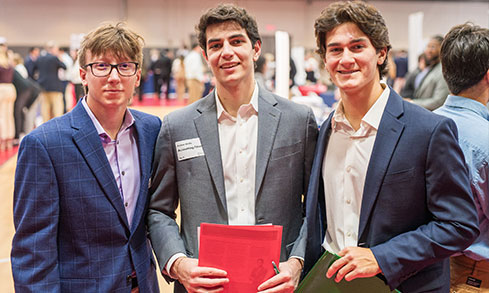 Three young men in suits and ties pose together at a formal event, smiling and looking confident.