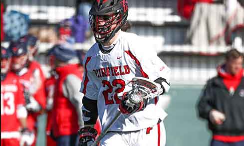 A lacrosse player in a white jersey with red accents that reads “Fairfield 22” holds a lacrosse stick. 