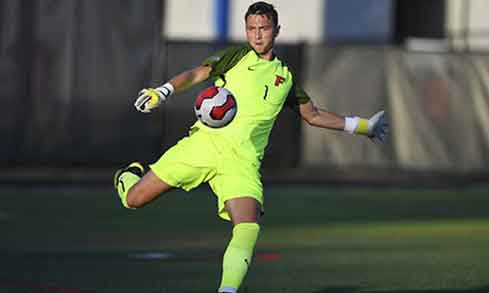 A goalie in a yellow jersey with a red “F” kicks a red and white soccer ball. 