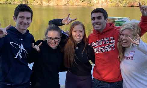 A group of students hold their hands up in a stag shape in front of a lake.