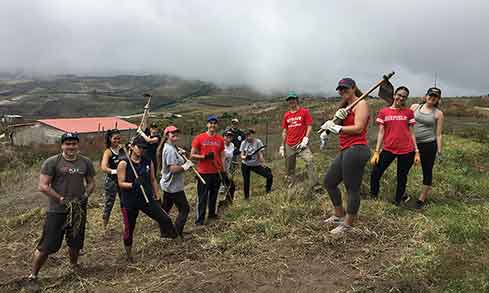 A group of students stand on a hill outdoors holding gardening tools.