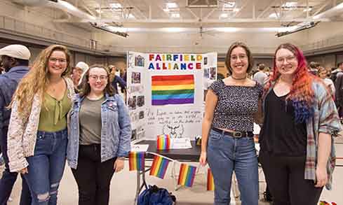 Four women stand beside a table displaying a vibrant rainbow flag, symbolizing diversity and inclusivity.