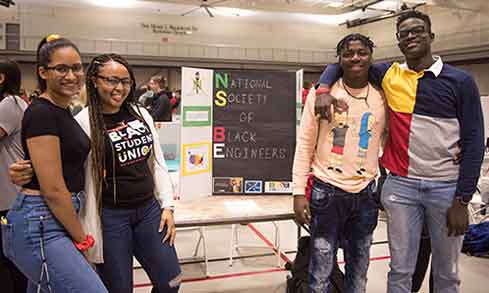 Four students smile together in front of a colorful poster board, capturing a fun moment in their school environment.