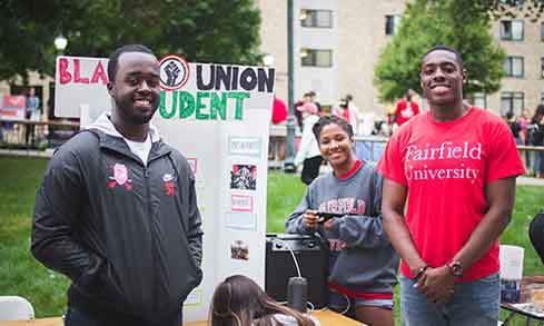 Three individuals positioned in front of a table displaying a sign, engaged in conversation or discussion.