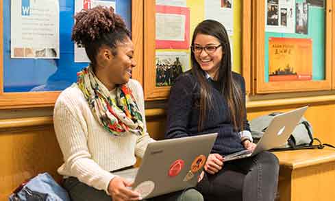 Two students seated on a bench in a library, engaged in study and discussion amidst bookshelves.