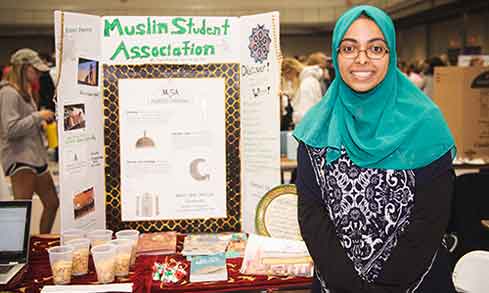 A woman in a hijab stands beside a table showcasing her display of items.