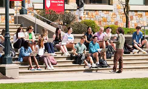 A class of students sit outdoors with laptops listening to their professor lecture.