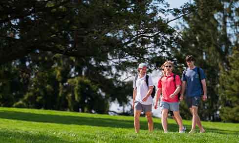 3 students walking by a grassy hill.