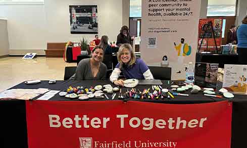 Two people sit at a table covered in pens that reads “Better Together: Fairfield University”.