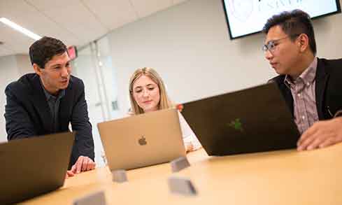 Two students and a professor talk with each other while working on laptops. 