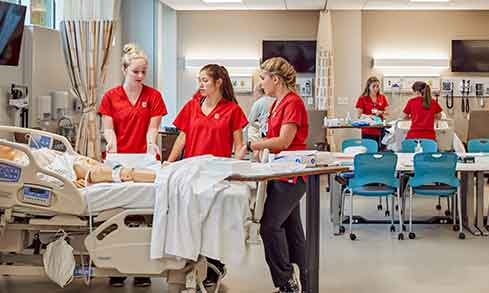 Three nursing students talk with each other while working with a dummy in a mock hospital room. 