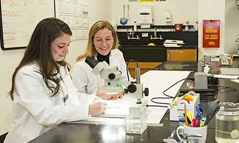 Two students in lab coats work a bench with a microscope. 