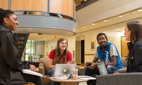 Four students sit at a table collaborating with laptops and notebooks.