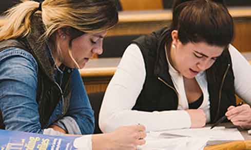 Two students sit at a desk in a classroom in front of textbooks and pieces of paper.