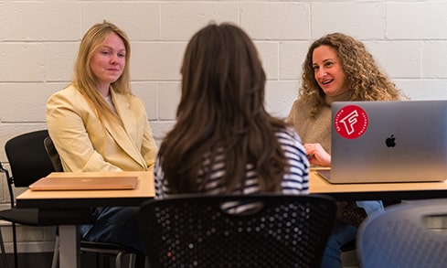 Two students sit at a table facing another student.