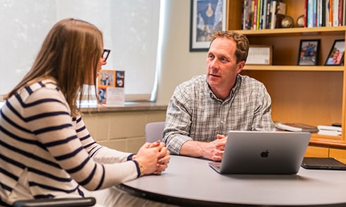 A student and professor sit in front of an open laptop and engage in conversation.