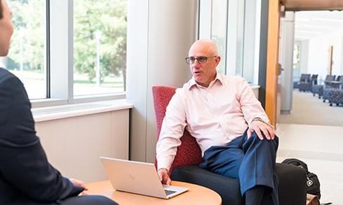 A professor sits in a chair while speaking to a student.