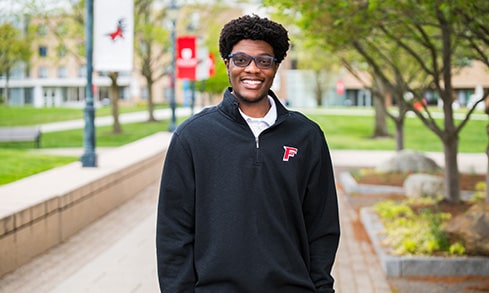 Male student with glasses smiling as he stands on a brick, campus walkway.