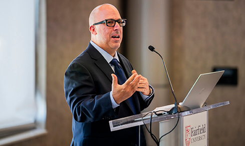 Gregg Caruso delivering a speech while standing at a Fairfield University podium with a laptop.