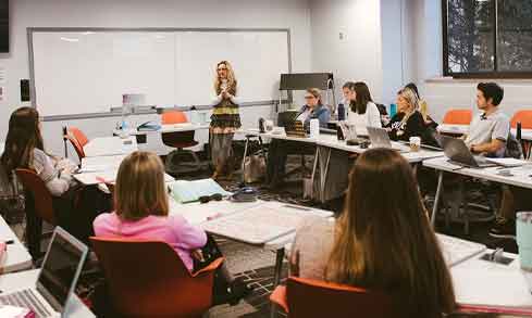 A classroom is full of students sitting in desks with open laptops in front of a standing professor. 