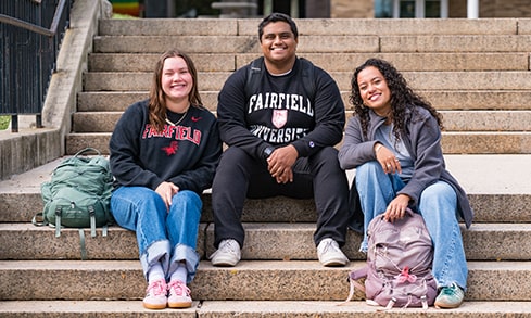 Three students in Fairfield apparel sitting and smiling on the steps to the Library.