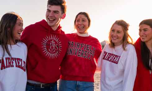 Five students wearing Fairfield merchandise laughing and posing for a photo on the beach. 